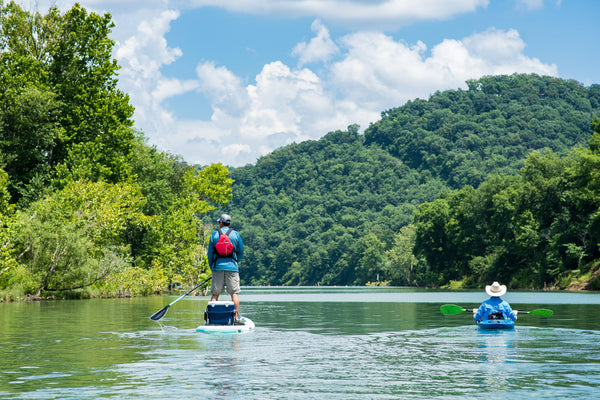 Man in blue shirt and white cowboy hat kayaks in a blue kayak next to a many in red pfd paddle boarding with a cooler on his paddle board.